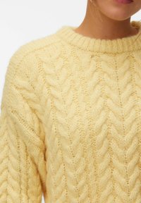 Gros plan d'une personne portant un pull en maille torsadée doux de couleur jaune pâle avec un col rond côtelé.