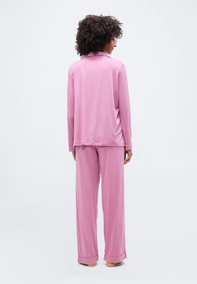 Γυναίκα με σγουρά μαλλιά που στέκεται, φορά χαλαρό ροζ μακρυμάνικο τοπ και παντελόνια με φαρδιά μπατζάκια, εμφανίζεται από πίσω σε λευκό φόντο.