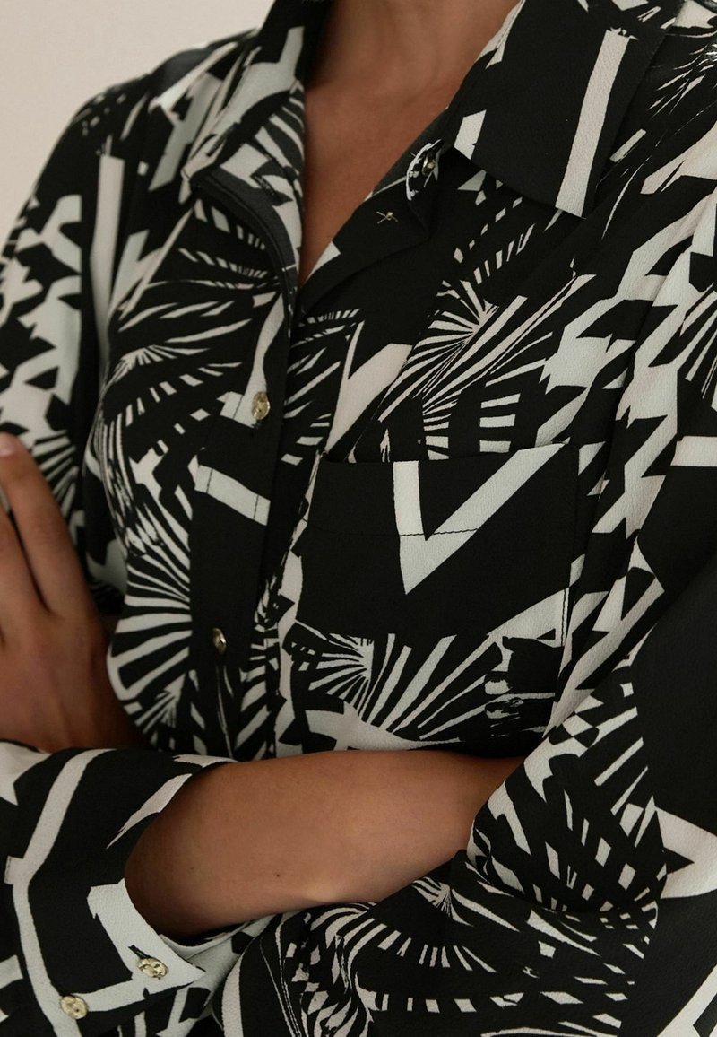 Chemise à boutons à motif noir et blanc, manches longues, poche avant, portée avec les bras croisés sur la poitrine.