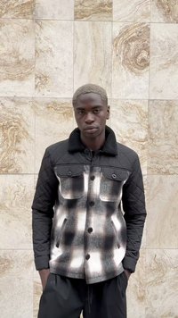 Mlad moški s kratkimi svetlimi lasmi, oblečen v črno-belo karirano jakno s podloženimi črnimi rokavi, stoji ob zidani steni iz ploščic.