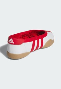 Bijeli tenisica s crvenim akcentima, s tri crvene pruge, glatkom završnom obradom i potplatom od gume. Logo je prikazan na peti.