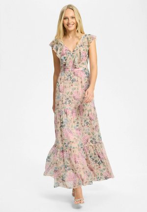 Mujer con un vestido largo sin mangas, con volantes y estampado floral, sonriendo y caminando hacia adelante sobre un fondo claro y liso.