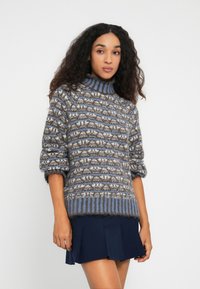 Kvinde med langt, krøllet hår iført en blå, grå og hvid mønstret sweater og en marineblå plisseret nederdel, stående foran en ensfarvet baggrund.