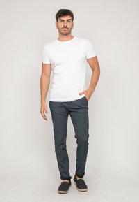 T-shirt blanc à manches courtes, pantalon à motifs sombres, espadrilles décontractées. Le modèle se tient avec une main dans la poche, sur fond neutre.