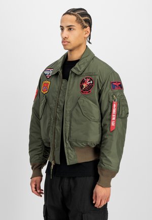 Junger Mann mit geflochtenem Haar, der eine olivgrüne Bomberjacke mit mehreren bunten Patches und einem roten Stoffetikett am Ärmel trägt.