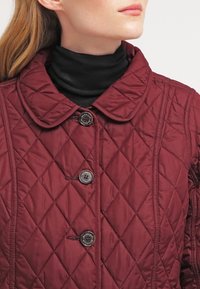 Gewatteerde bordeauxrode jas met een ronde kraag, voorzien van twee zwarte knopen en diagonale stiksels. Gedragen over een zwarte coltrui.