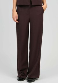 Personne portant un pantalon large marron foncé, se tenant avec une main dans la poche et des chaussures noires brillantes sur un fond gris.