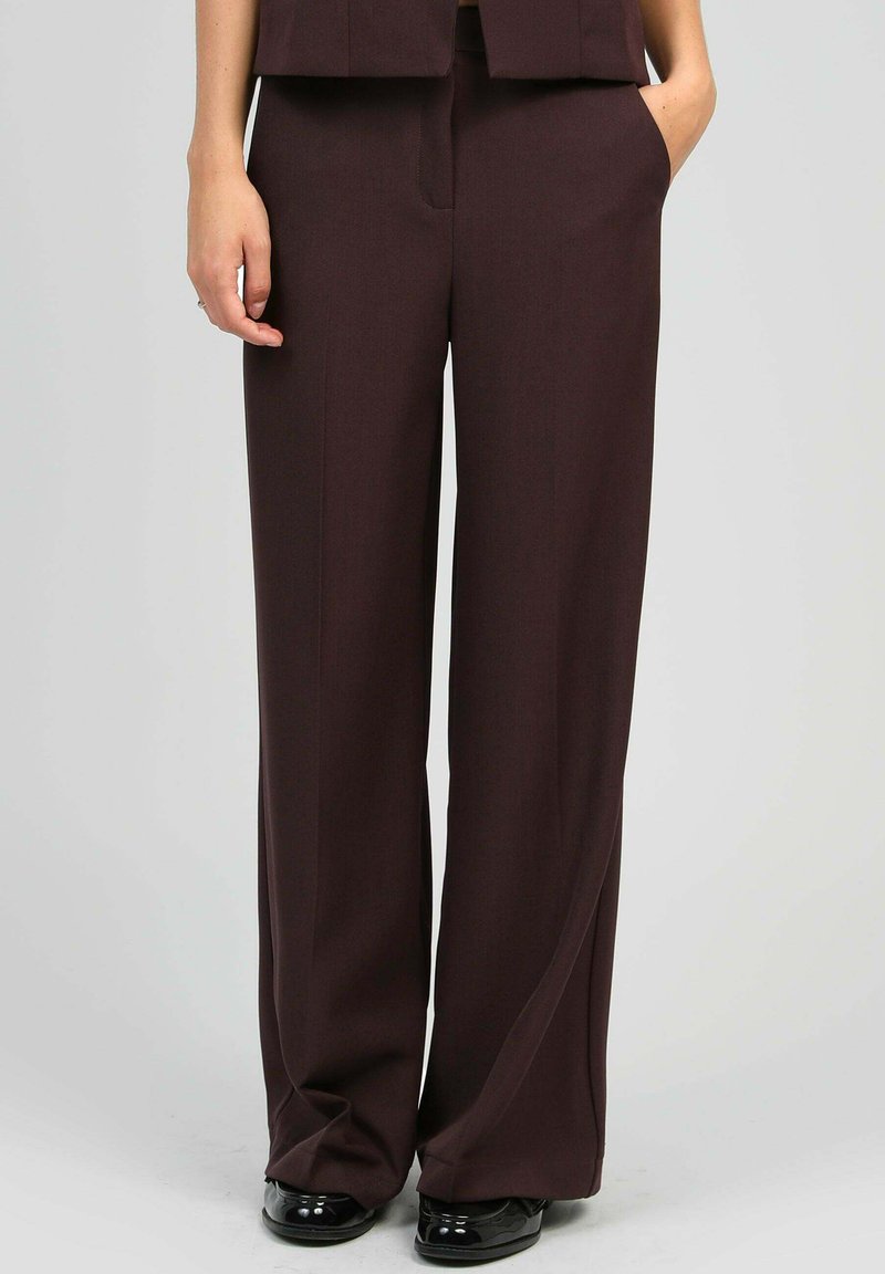 Personne portant un pantalon large marron foncé, se tenant avec une main dans la poche et des chaussures noires brillantes sur un fond gris.