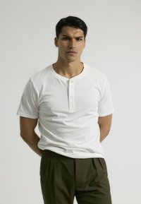 Camisa henley de manga corta blanca con tapeta de tres botones, fabricada en algodón suave, combinada con pantalones con pliegues de color verde oliva. Diseño simple.
