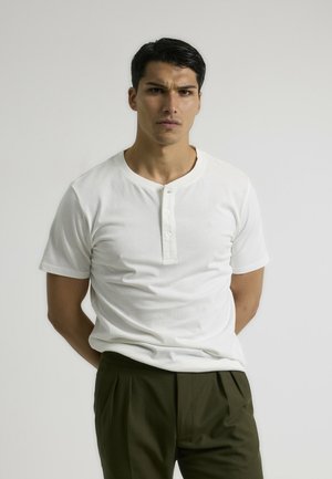 Camisa henley de manga corta blanca con tapeta de tres botones, fabricada en algodón suave, combinada con pantalones con pliegues de color verde oliva. Diseño simple.