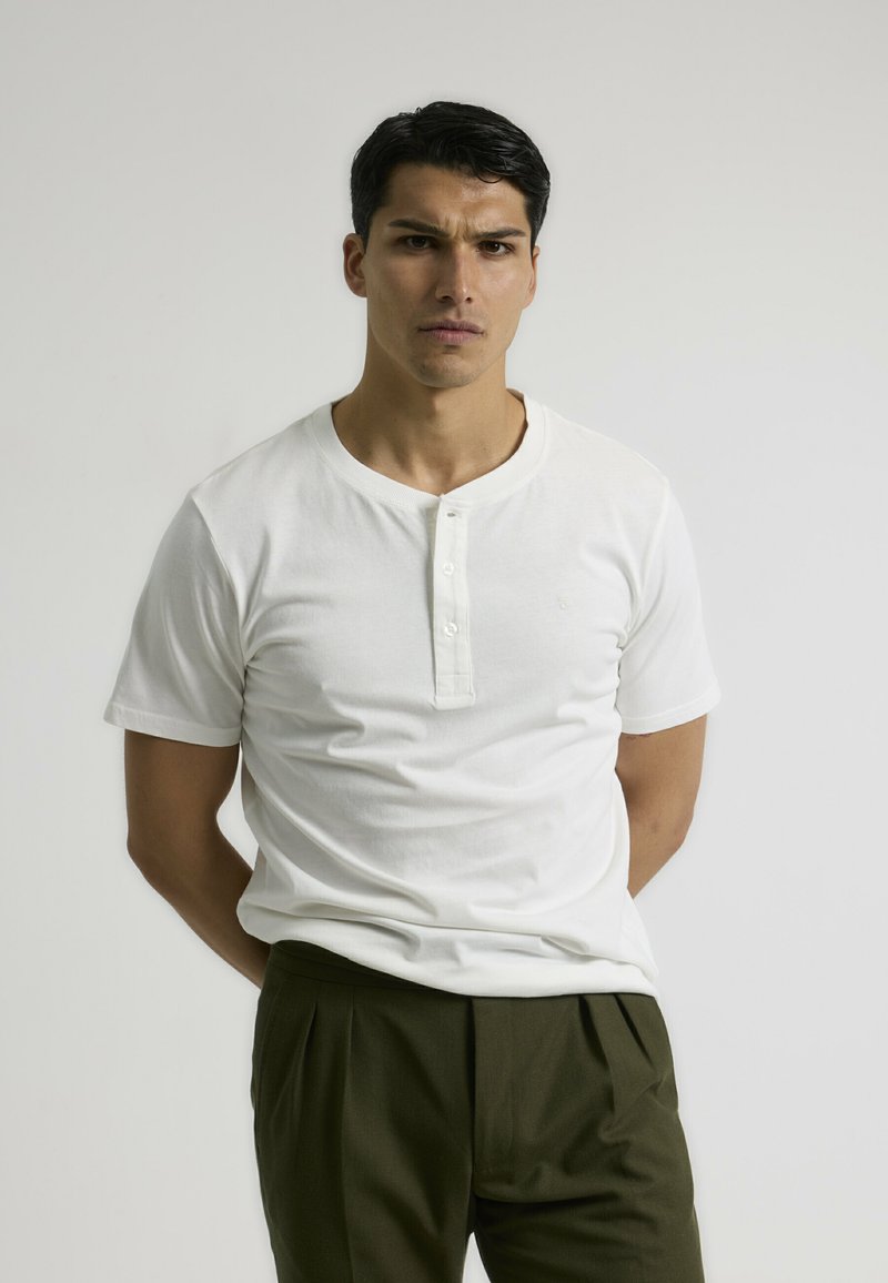 Camisa henley de manga corta blanca con tapeta de tres botones, fabricada en algodón suave, combinada con pantalones con pliegues de color verde oliva. Diseño simple.
