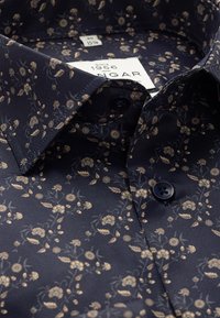 Marineblaue Bluse mit floralem Muster in kunstvollen Gold- und hellbraunen Farbtönen. Verfügt über einen spitzen Kragen und dunkle Knöpfe. Hergestellt aus glattem Stoff.