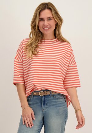 Vrouw glimlacht, draagt een koraalrood-wit gestreept T-shirt met korte mouwen, blauwe spijkerbroek met een beige riem en gouden oorringen.