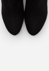 Bottes cheville en daim noir avec un bout pointu et un détail de couture, présentant une texture lisse et des éléments de design minimalistes.