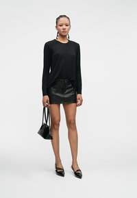 Top negro de manga larga con detalles fruncidos, combinado con una minifalda ajustada de cuero negro y tacones negros de punta, sosteniendo un pequeño bolso negro.