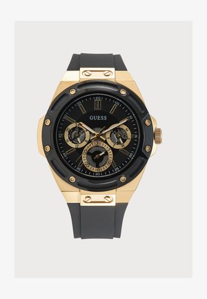 Montre bracelet Guess noire et dorée avec cadran rond, trois sous-cadrans, index dorés et bracelet noir avec vis dorées sur le boîtier.