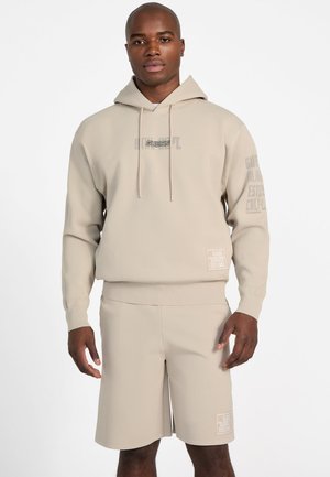 Mand iført beige hættetrøje med "GUESS" logo og matchende beige shorts med logopatches, stående mod en hvid baggrund.