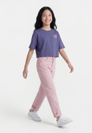 Κορίτσι που περπατά, φοράει μωβ κοντό T-shirt, ανοιχτό ροζ παντελόνι γυρισμένο προς τα πάνω και άσπρα αθλητικά παπούτσια, χαμογελώντας μπροστά σε απλό λευκό φόντο.