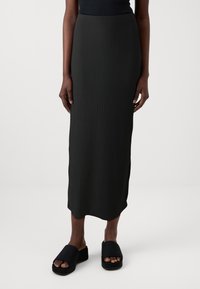 Schwarzer gerippter Midirock mit tailliertem Bund und seitlichen Schlitzen, kombiniert mit schwarzen Plateausandalen mit breiten Riemen. Glatte Textur überall.