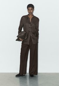 Kobieta w luźnej, ciemnobrązowej koszuli zapinanej na guziki i szerokich spodniach trzyma fakturowaną, ciemnobrązową kopertówkę, stojąc na tle jasnej, gładkiej ściany.