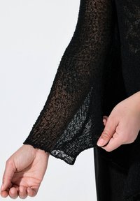 Svart, texturerad stickad tröjsleeve med lös passform och fransad kant. Materialet verkar lätt och halv genomskinligt. Händer som håller i tyget.