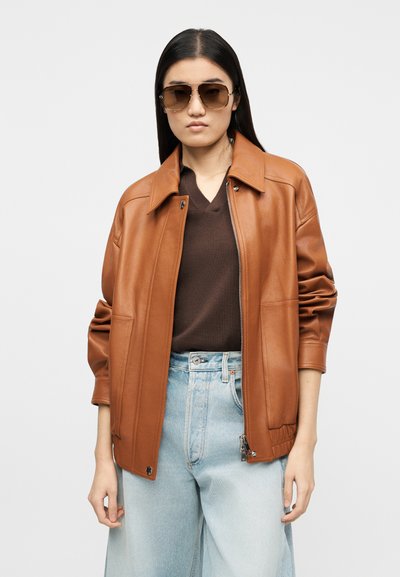 Νεαρό άτομο που φορά καφέ δερμάτινο μπουφάν, σκουρό καφέ πουλόβερ με V λαιμόκοψη, ανοιχτό μπλε τζιν και μεγάλα καφέ γυαλιά ηλίου, στέκεται μπροστά σε λευκό φόντο.