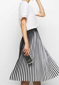 Femme portant un chemisier blanc et une jupe plissée à rayures verticales noir et blanc, tenant une pochette argentée réfléchissante.