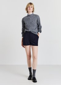 Grauer Strickpullover mit Perlenakzenten, kombiniert mit dunkelblauen Shorts. Das Model trägt graue Socken und schwarze Slipper. Minimalistischer Hintergrund.