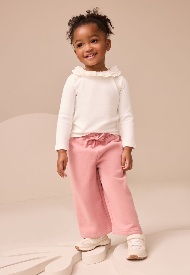 Haut blanc à manches longues avec un col à volants, associé à un pantalon de jogging ample rose clair. L'enfant porte des baskets blanches à scratchs.