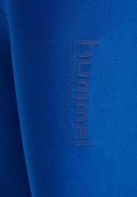 Blauwe prestatieleggings van een gestructureerd, rekbaar materiaal met een subtiele glans en een verhoogd logo in donkerblauw langs de zijkant.