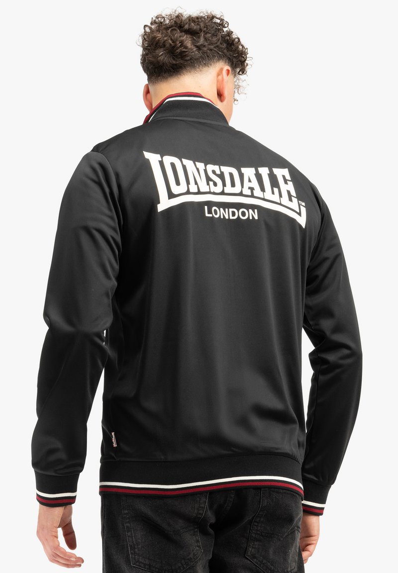 Zwarte bomberjack met witte "LONSDALE" letters en "LONDON" eronder, voorzien van een geribde kraag en zoom met rode en witte strepen.