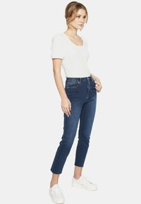 Biała bluzka z krótkim rękawem i okrągłym dekoltem, zestawiona z ciemnoniebieskimi jeansami z wysokim stanem. Model nosi białe sneakersy. Minimalistyczny design i krój.