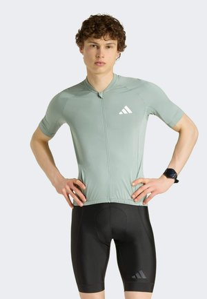 Ung mand med krøllet hår iført en tætsiddende grå cykeltrøje og sorte polstrede shorts, hænderne på hoften, foran en ensfarvet baggrund.