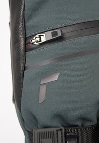 Grüner Stoff-Rucksack mit einem schwarzen Lederakzent, ausgestattet mit einem Reißverschluss mit metallischen Beschlägen und einem grauen Logo auf der Vorderseite.