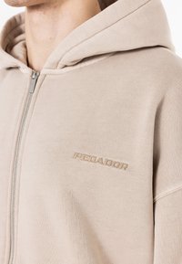 Beige zip-up hættejakke lavet af struktureret stof, med et broderet logo på brystet og en snor i hætten.