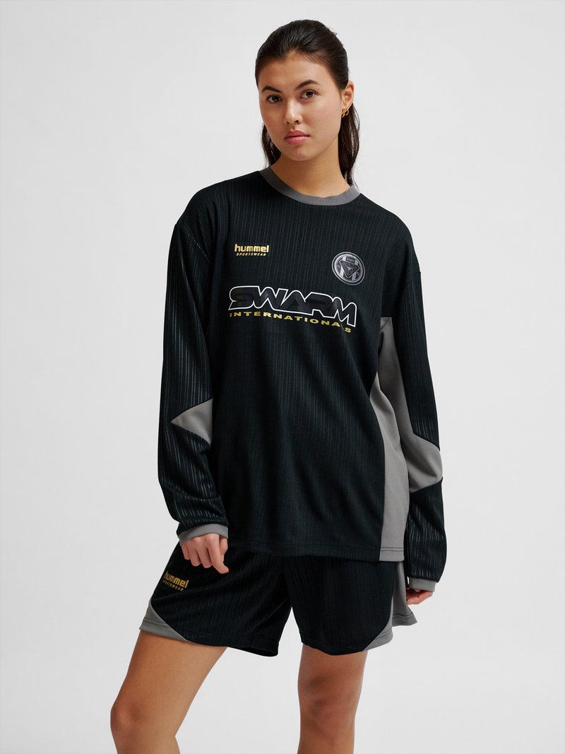 Junge Frau trägt ein schwarz-graues langärmliges Sporttrikot und passende Shorts mit dem Logo "SWARM INTERNATIONALS" und steht vor einem einfarbigen Hintergrund.