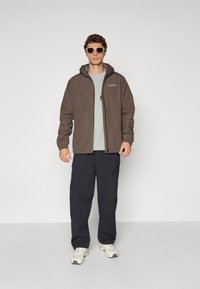 Bărbat îmbrăcat într-o jachetă cu glugă maro, cămașă gri, pantaloni cargo negri, adidași albi și ochelari de soare, stând pe un fundal simplu deschis.