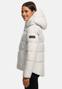 Veste doudoune gris clair avec capuche, texture matelassée, fermeture éclair sur le côté et accent logo sur la manche, offrant un design moderne et décontracté.