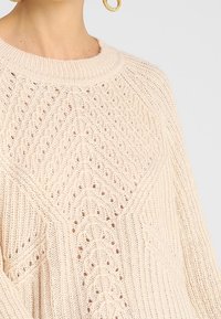 Krémový pletený svetr s kulatým výstřihem, texturovaným diamantovým vzorem a otevřeným tkaním. Obsahuje spadlá ramena a lehký materiál.