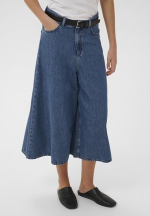 Osoba u širokim skraćenim plavim trapericama, crnim remenom, bijelom košuljom i crnim cipelicama bez vezica stoji na jednobojnoj pozadini.