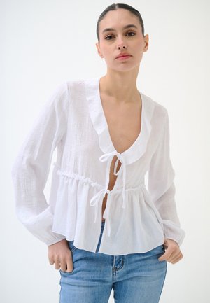 Donna che indossa una camicetta bianca strutturata con colletto arricciato e lacci, abbinata a jeans azzurri chiari su sfondo bianco semplice.