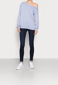 Sudadera azul claro off-shoulder con mangas largas y puños acanalados, combinada con jeans ajustados oscuros y zapatillas blancas sobre un fondo liso.