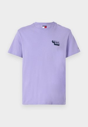 Camiseta de algodón lavanda con cuello redondo, mangas cortas y un logotipo negro de "TOMMY JEANS" en el pecho. Textura de tela suave.