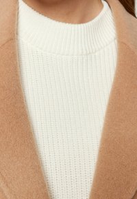 Cappotto di lana marrone chiaro con una texture morbida e liscia, sovrapposto a un maglione bianco a costine. Primo piano sul colletto e sui dettagli del tessuto.