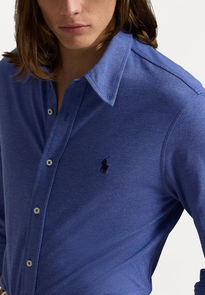 Camisa de polo de mangas longas azul, feita de um tecido texturizado. Apresenta um colarinho com botões, uma pala frontal com botões e um pequeno logotipo bordado no peito.