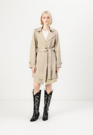 Beige trench coat with a belt, buttoned cuffs, and wide lapels, paired with black cowboy boots featuring silver embroidery.