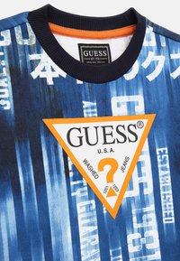 Felpa grafica blu con un audace patch triangolare arancione con il testo "GUESS". Il materiale sembra morbido; il design include motivi a strisce sovrapposti.