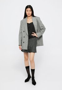 Blazer gris croisé à rayures verticales, associé à un haut noir et une jupe plissée grise texturée. Des chaussettes noires et des chaussures pointues complètent le look.