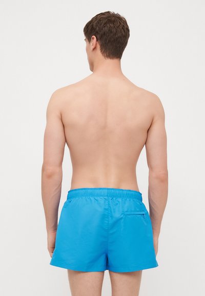 Un short de bain bleu avec une ceinture élastique et une poche arrière. Le tissu léger a une texture lisse et une couleur unie.