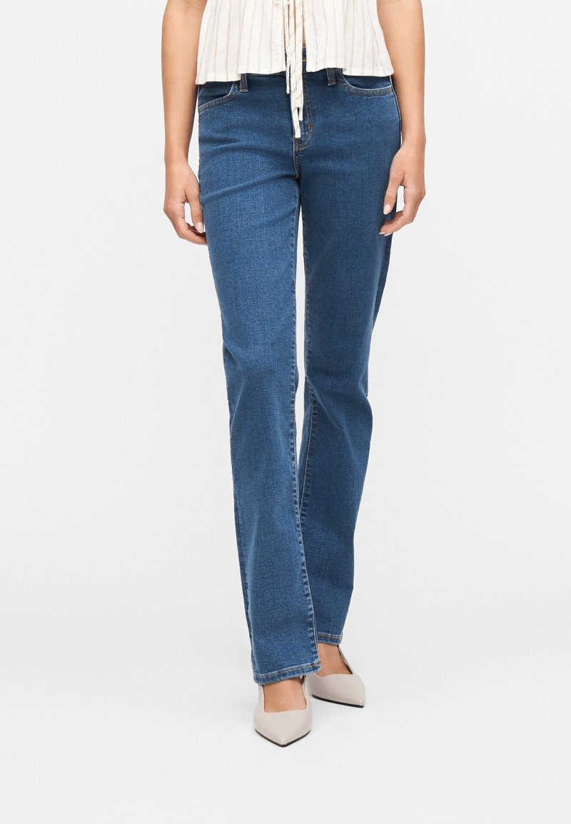 Femme portant un jean bleu droit et des chaussures blanches à bouts pointus, debout devant un fond clair uni.